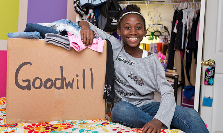 a young girl sits on the bed in her room next to a box of donations for Goodwill