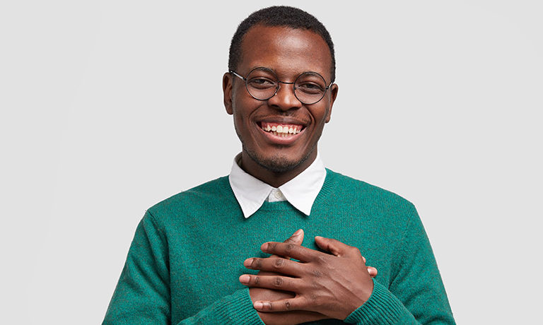 a young man wearing glasses and a green sweater puts his hands over his heart in a "thank you" gesture