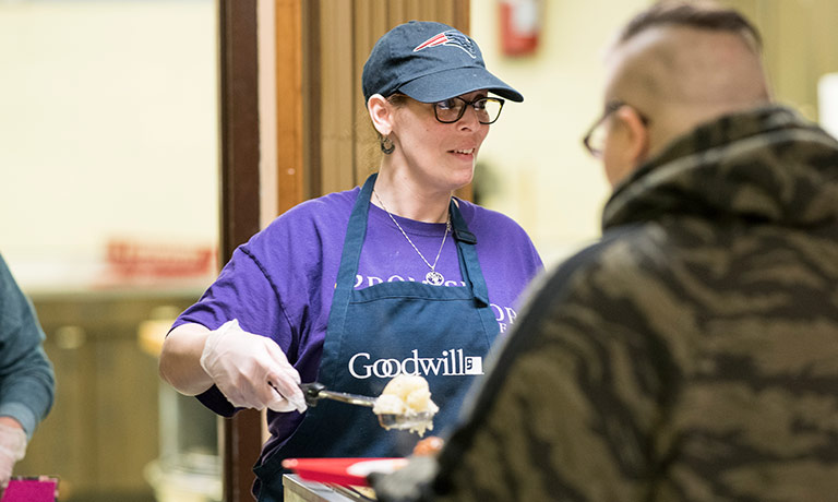 a woman wearing a Goodwill apron serves food to a man at a soup kitchen